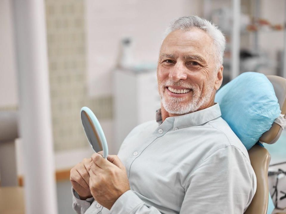 A man is sitting in a dental chair looking at his teeth in a mirror.