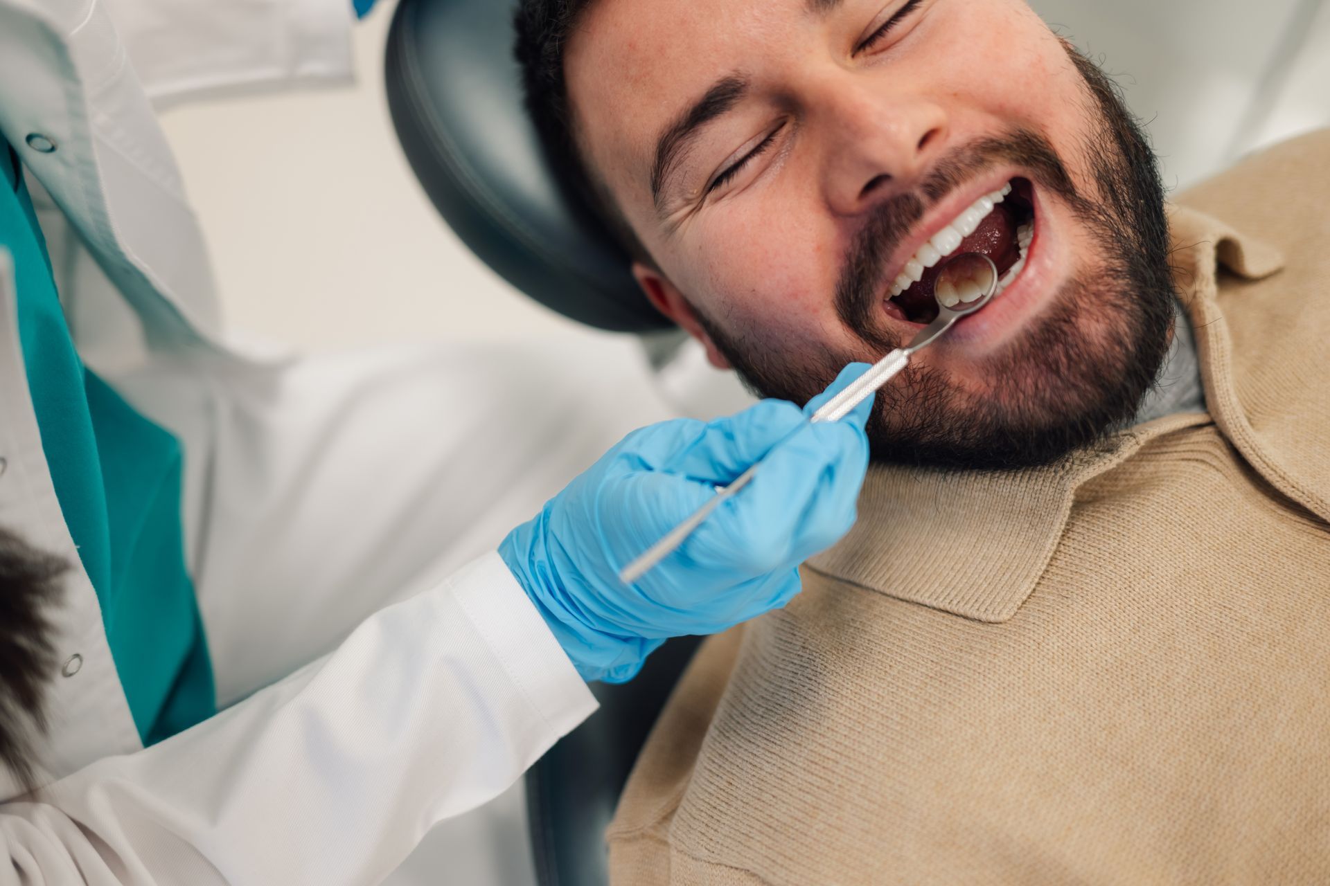 A dental professional in blue gloves uses a dental mirror to examine the teeth of a person sitting in a dental chair.