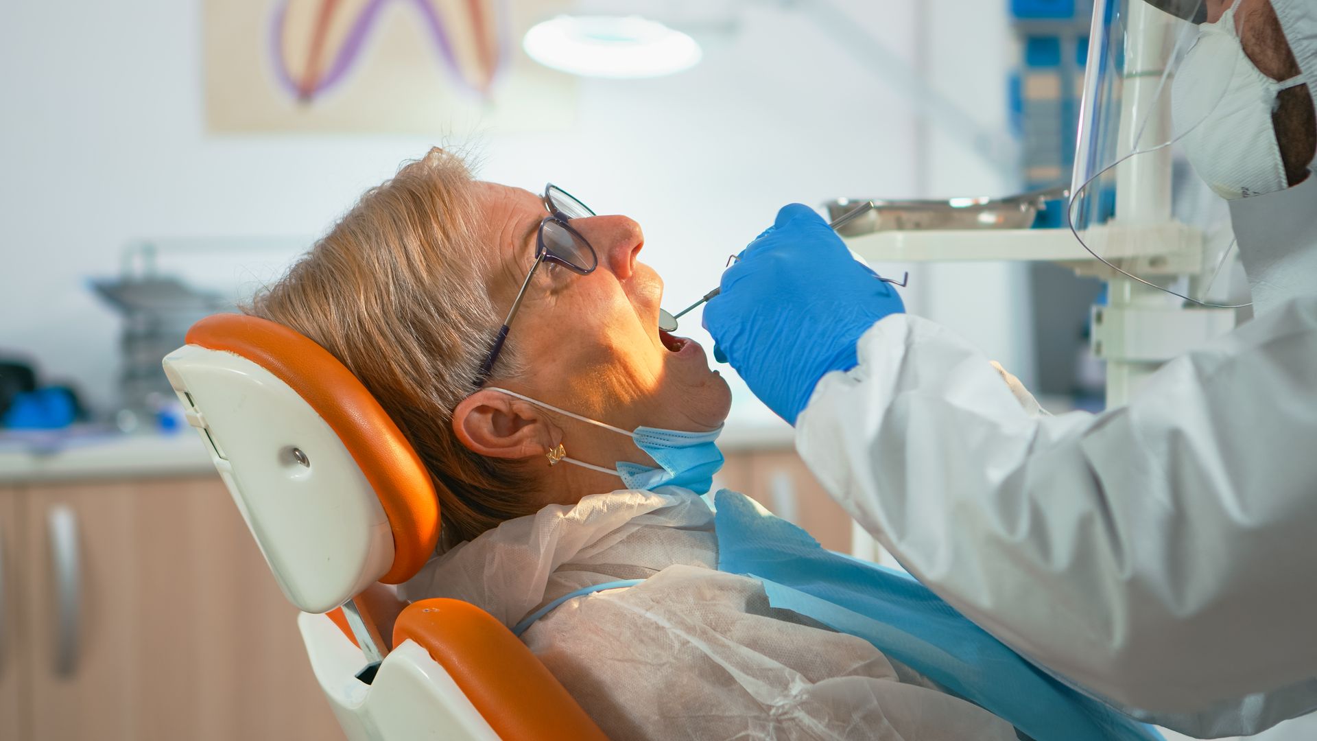 Patient in dentist chair, mouth open, dentist in protective gear examining teeth with tools.