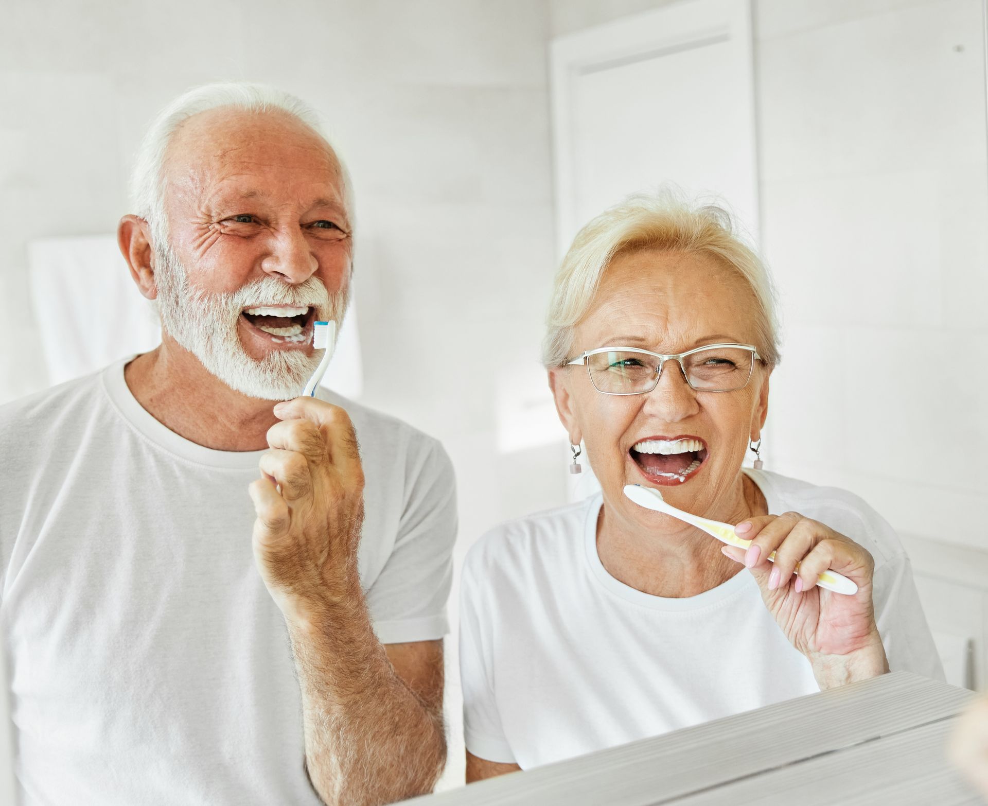 Two smiling people brushing their teeth in a bright bathroom mirror.