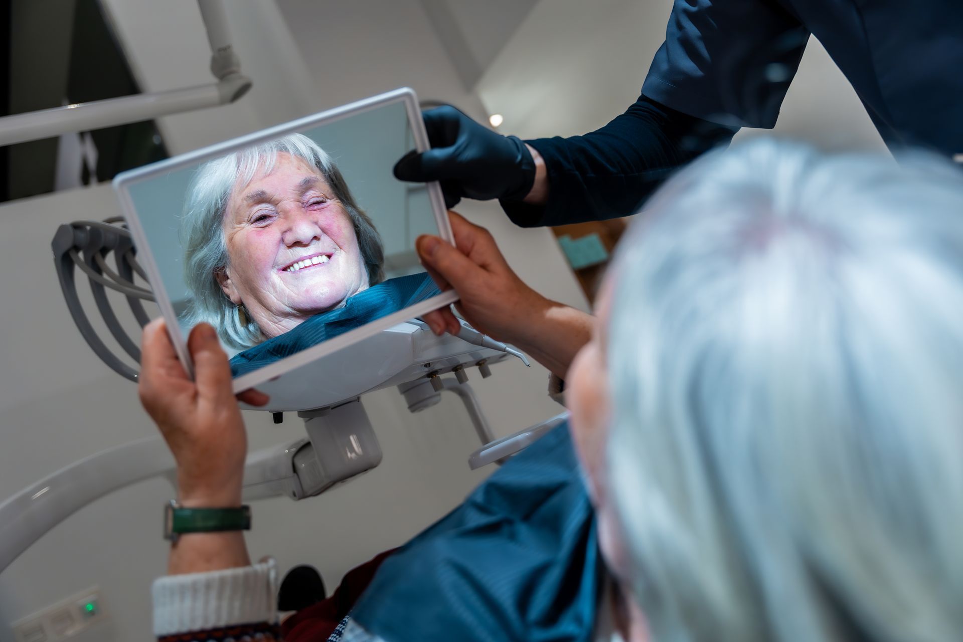 A dental patient holds a mirror to view their smile while a professional wearing gloves assists them.