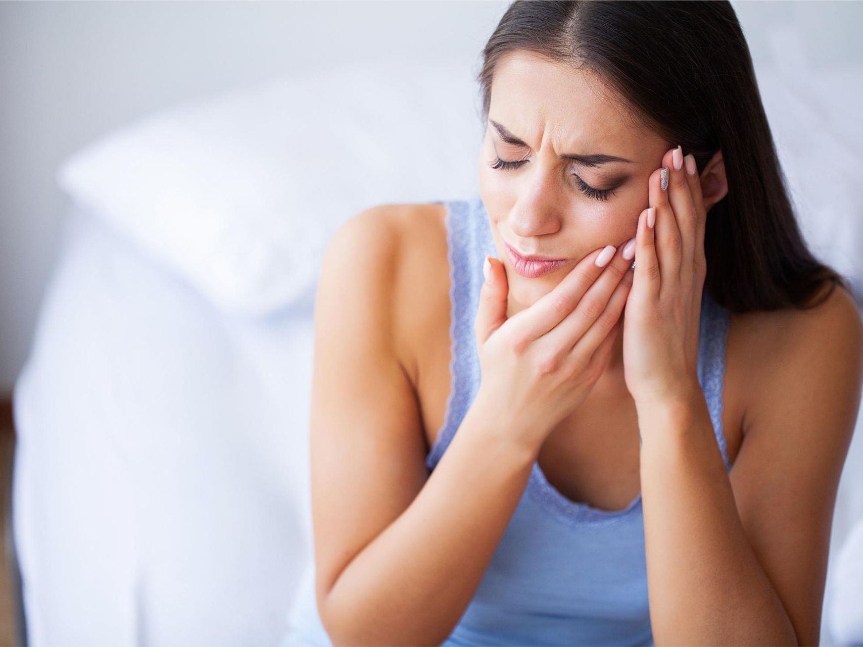 A woman is sitting on a bed holding her face in pain.