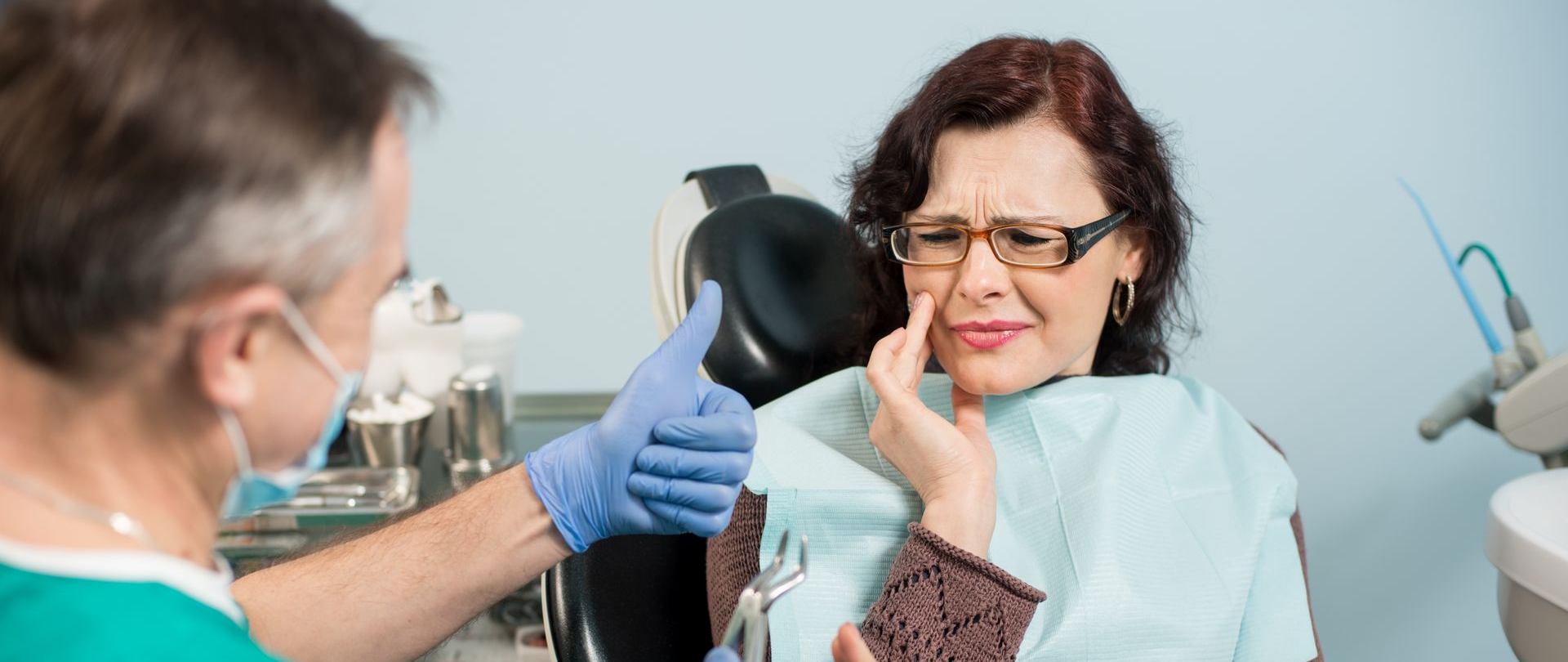 Dentist examining a patient with a toothache in a dental office. The patient is grimacing and holding her face.