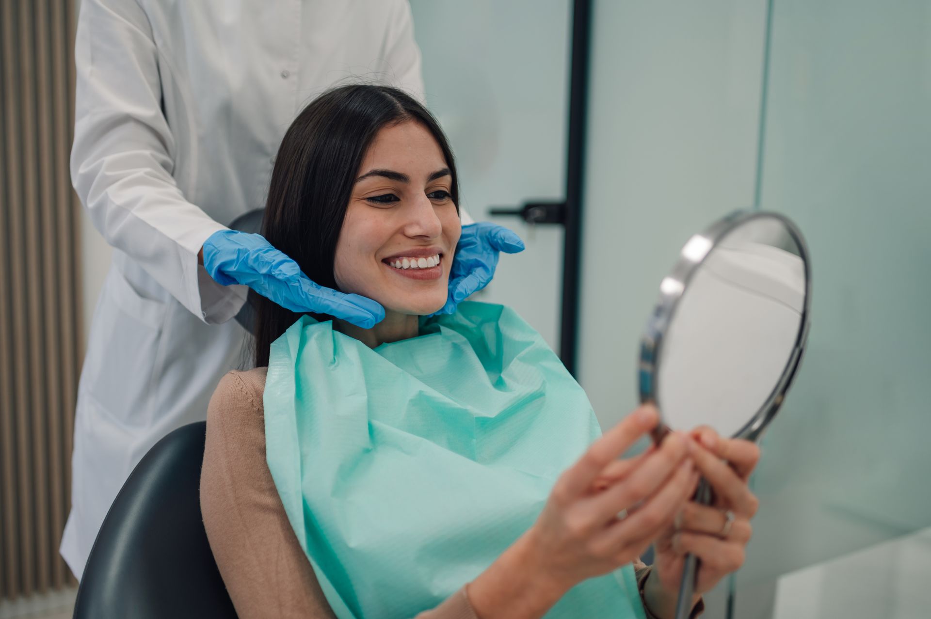 Woman smiles at her teeth in a mirror, a dentist in gloves checks her face in a dental office.