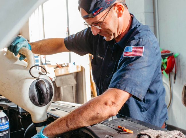 Mechanic pouring fluid into a car engine, wearing a cap, glasses, and blue gloves.