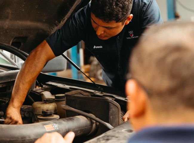 Mechanic working on a car engine in a workshop, another person observing.