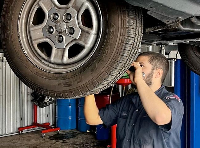 Mechanic inspecting a car's underside, holding a light. Garage setting, blue lift, tire in focus.