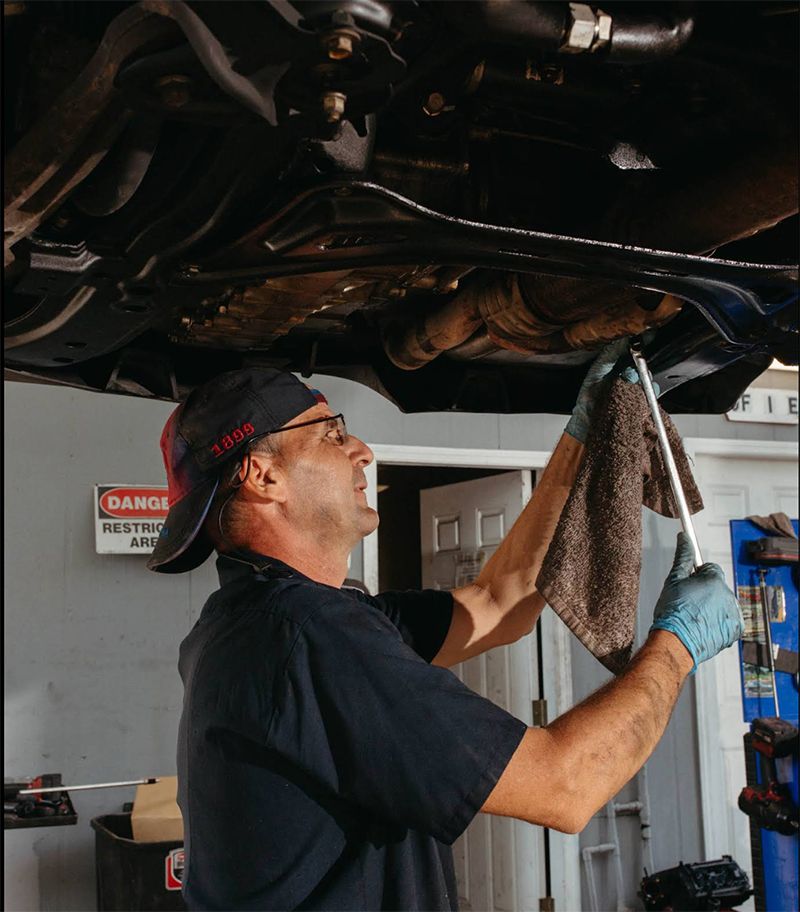 Mechanic working under a car, wiping with a cloth. Garage setting, wearing cap and gloves.