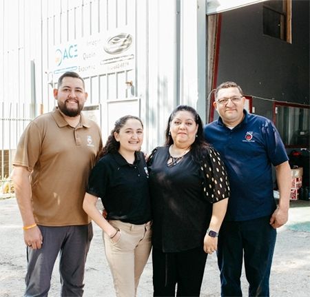 Four people smiling in front of a business with a sign. Brown and black attire, outdoor setting.