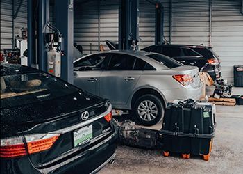 Cars in a repair shop; silver sedan on a lift, a black Lexus, and equipment in the foreground.