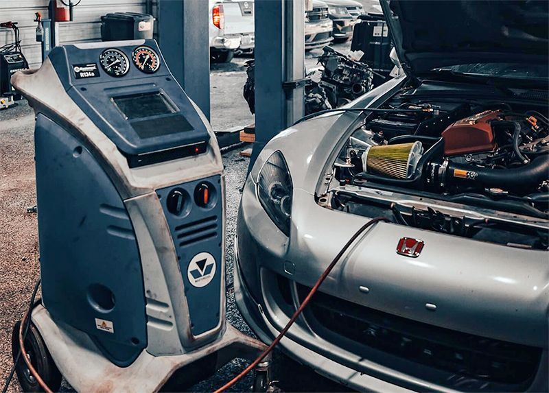 A silver car with its hood open is hooked up to a blue machine in an auto repair shop.