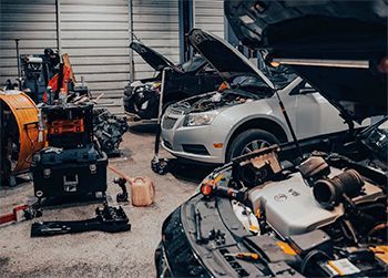 Cars with open hoods inside a garage; tools and equipment are scattered around.