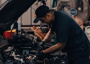 Mechanic working on a car engine in a garage. He wears a black cap, shirt, and gloves.
