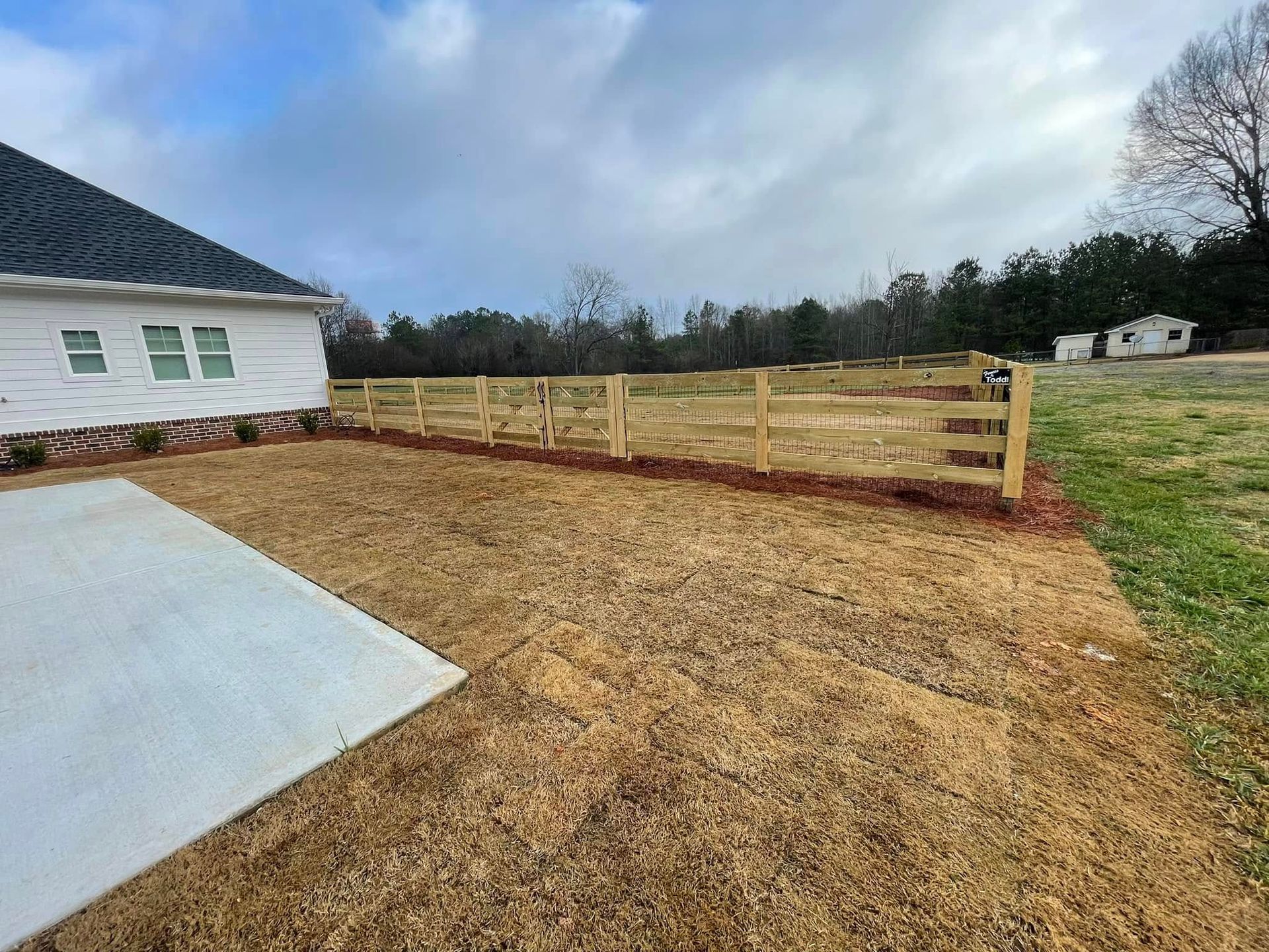 Freshly graded dirt yard beside a white house, with a concrete slab and wooden fence under cloudy sky