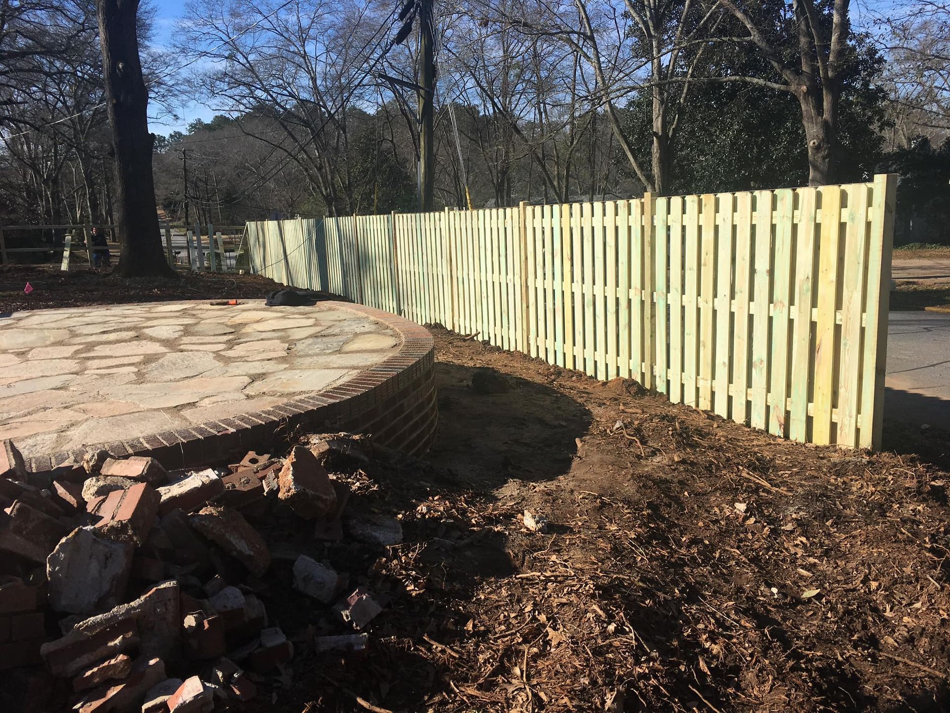 Freshly cut tree stump beside stacked firewood and a pale wooden fence in a wooded yard