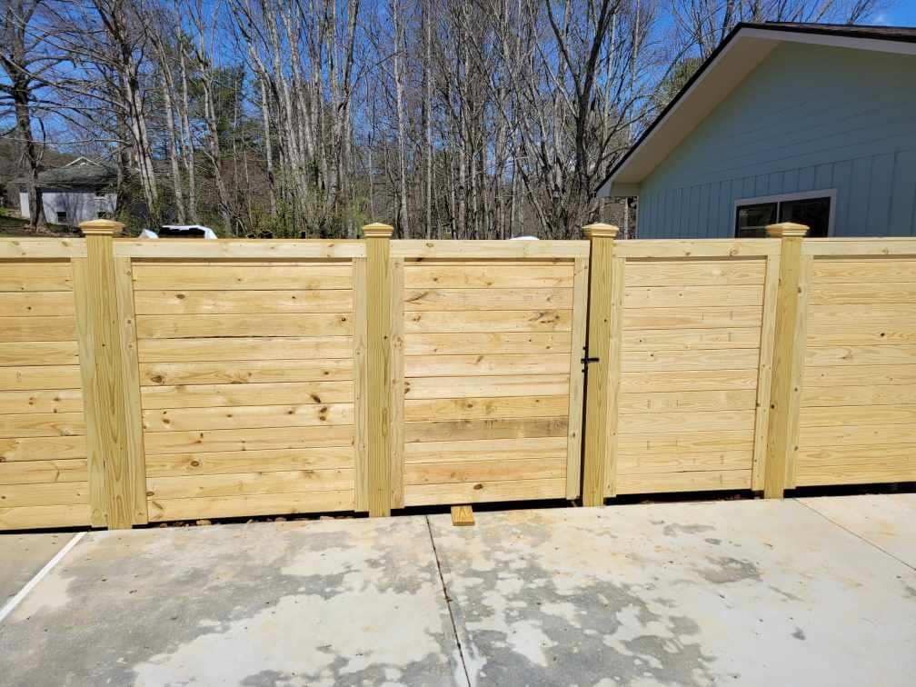 Wooden privacy fence with gates along a concrete driveway beside a house and trees