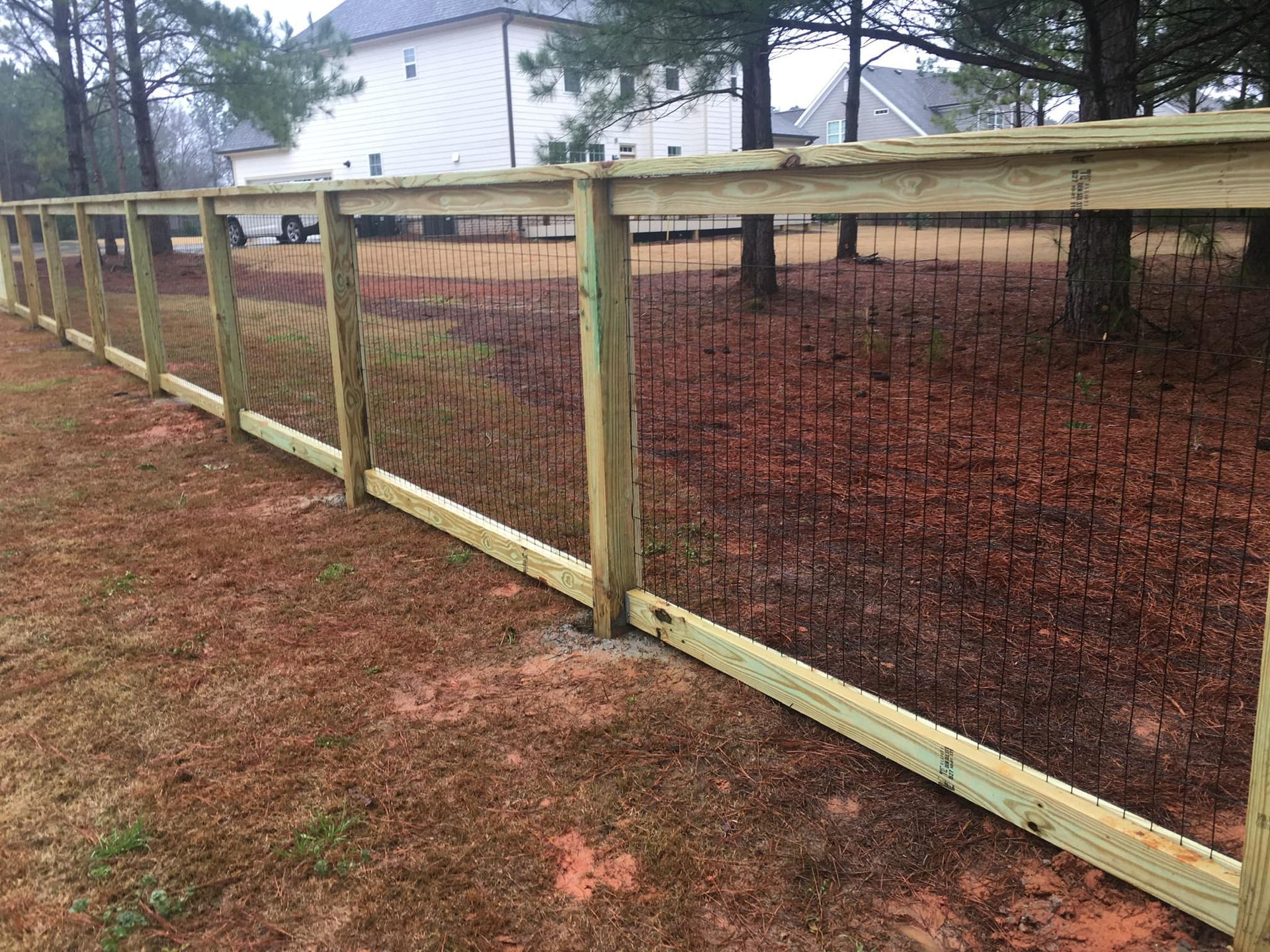 Wooden fence with black mesh panels along a dirt yard beside a white building and trees