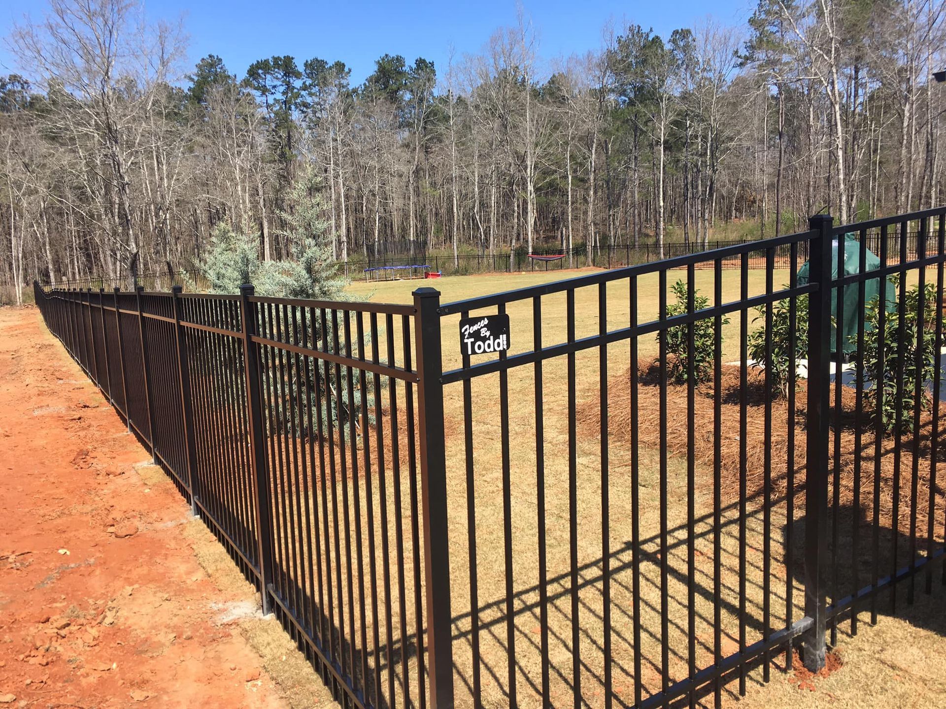 Black metal gate beside a red dirt path in a wooded outdoor area