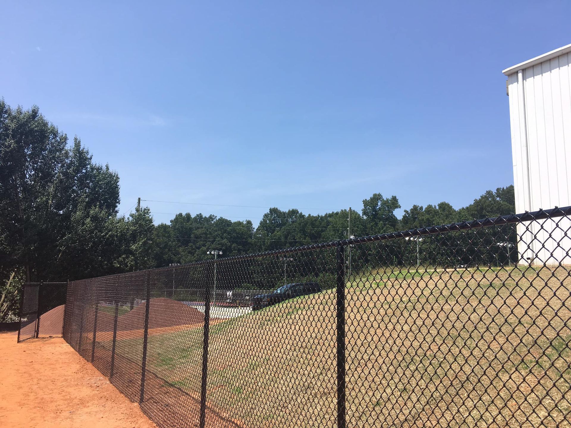 Chain-link fence beside a dry grassy field under a clear blue sky, with trees and a white building at right