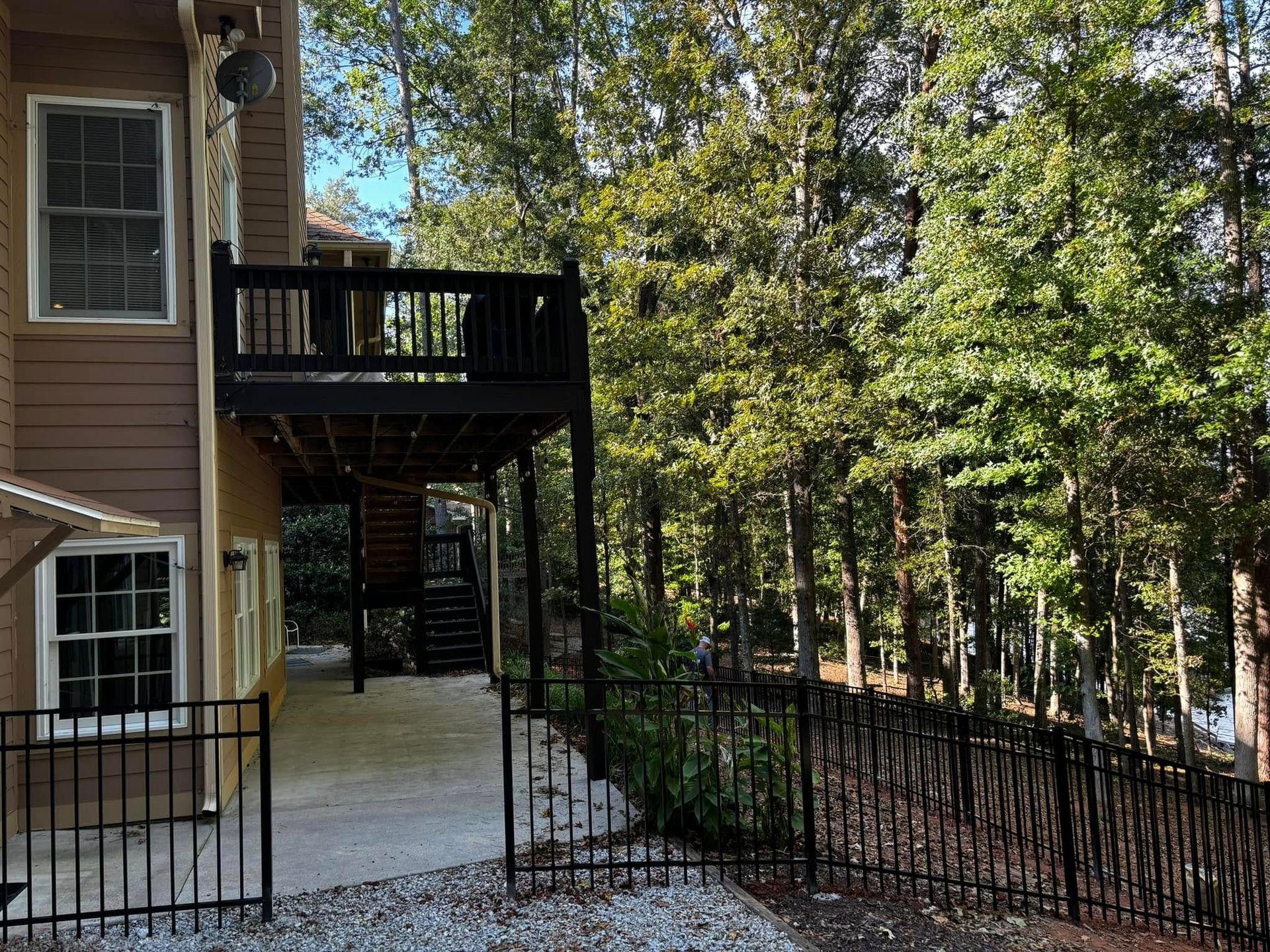 Apartment building balcony and walkway beside a wooded area, with black metal railings and trees in the background