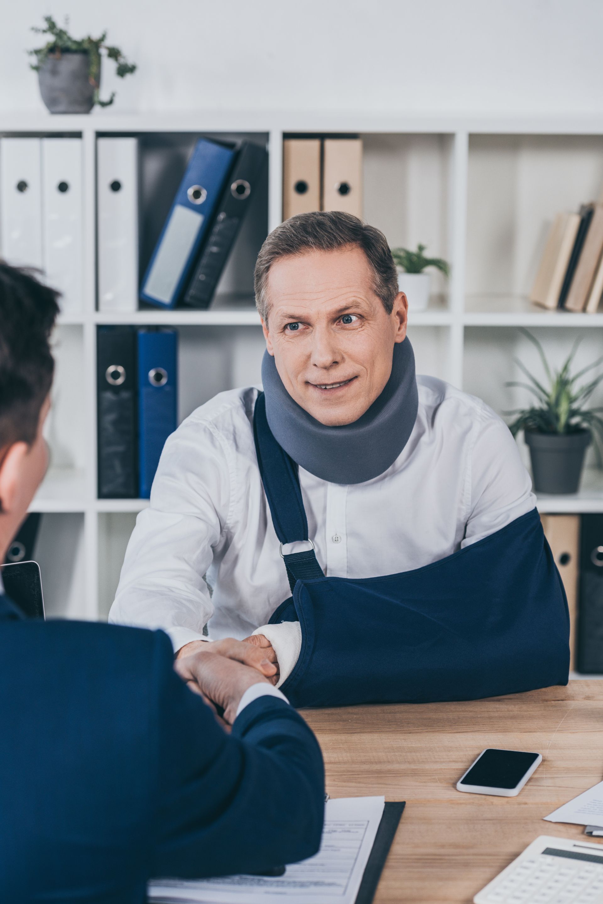 A professional wearing a neck brace and arm sling shaking hands with an unseen colleague in an office.