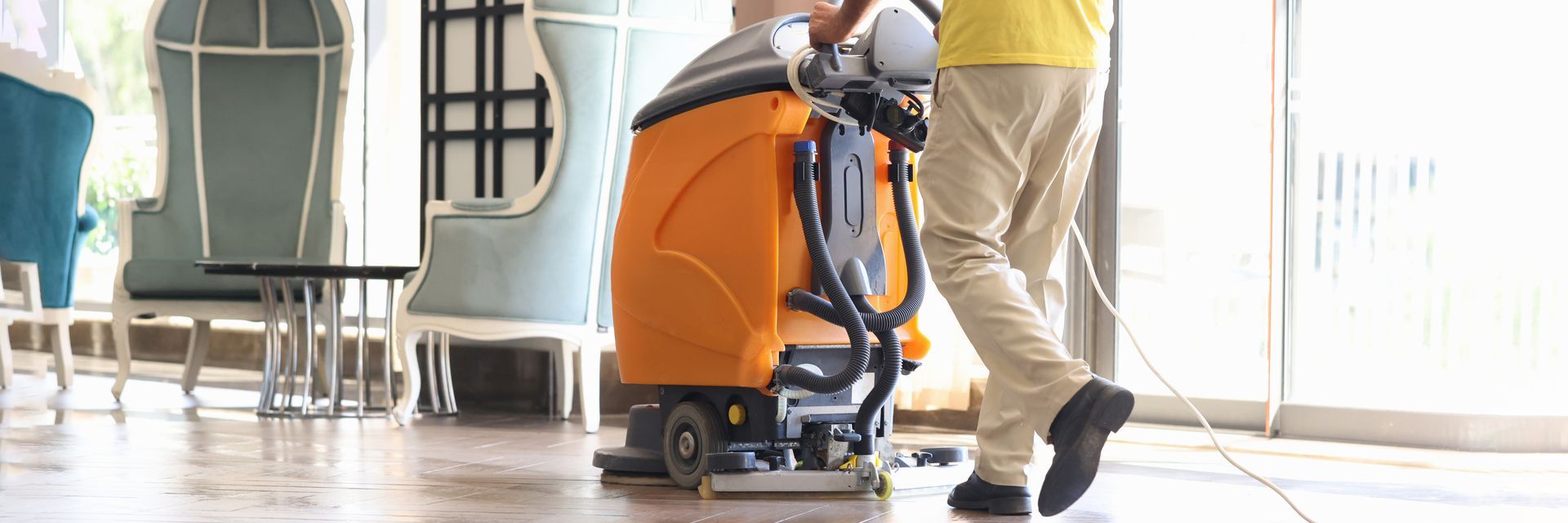 A person cleaning a glossy floor with an orange floor-cleaning machine indoors, near a window.