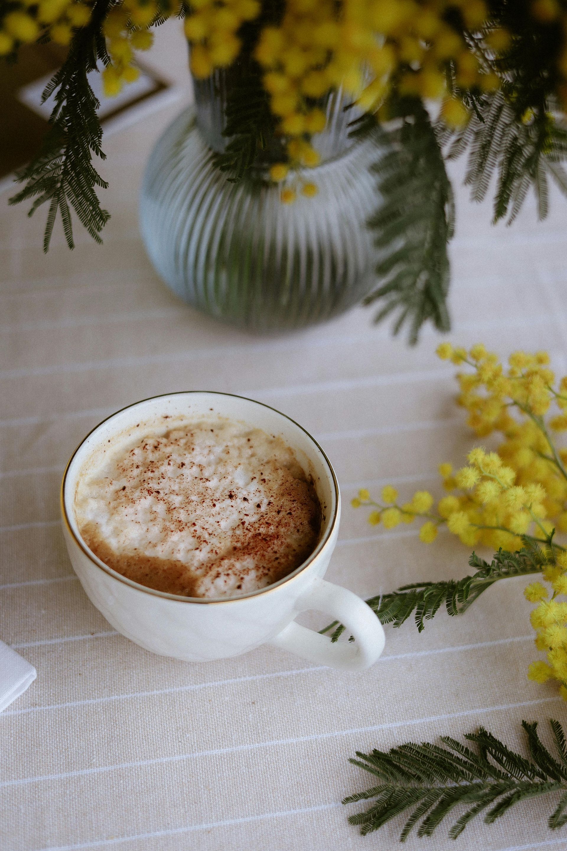 A cup of cappuccino on a white tablecloth with yellow mimosa flowers and a blue vase in the background.