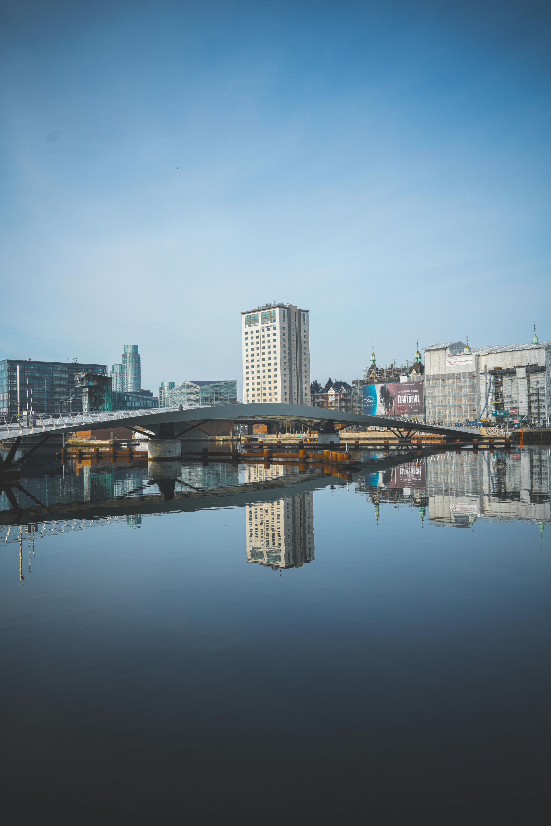 City skyline reflected in calm water under a blue sky. Buildings and a bridge are visible.