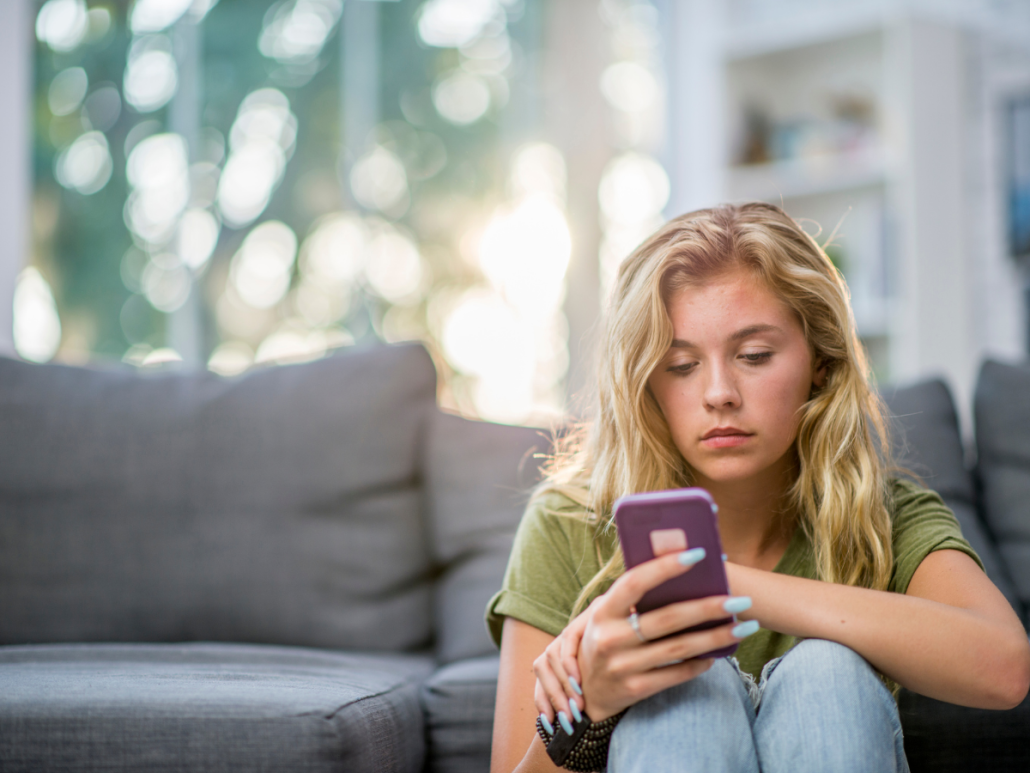 Hope Against Trafficking | A young woman with long blonde hair sits on the floor next to a couch, looking at her smartphone with a serious expression.