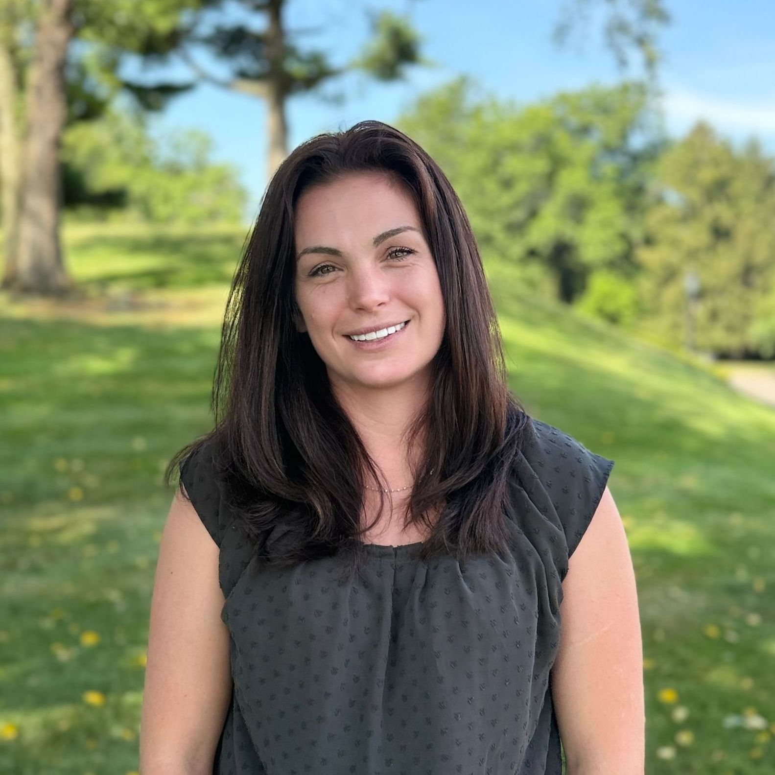 A woman in a gray shirt is smiling for the camera in a park.