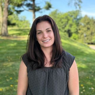 A woman in a gray shirt is smiling for the camera in a park.