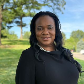 A woman in a black dress and pearls is smiling for the camera in a park.