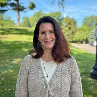 A woman wearing a sweater and a necklace is smiling for the camera in a park.