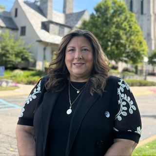 A woman wearing a black jacket and a necklace is standing in front of a building.