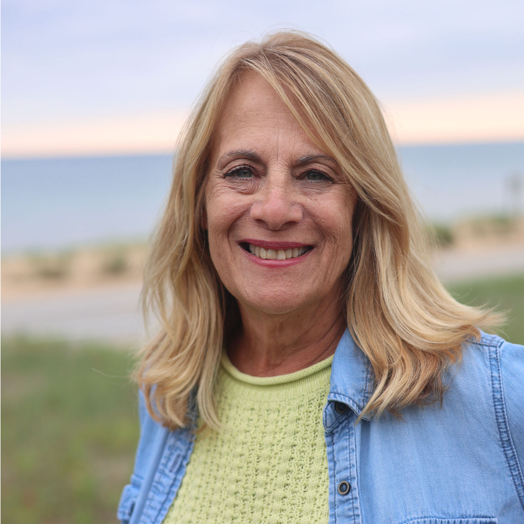 A woman wearing a denim jacket and a yellow sweater is smiling for the camera.
