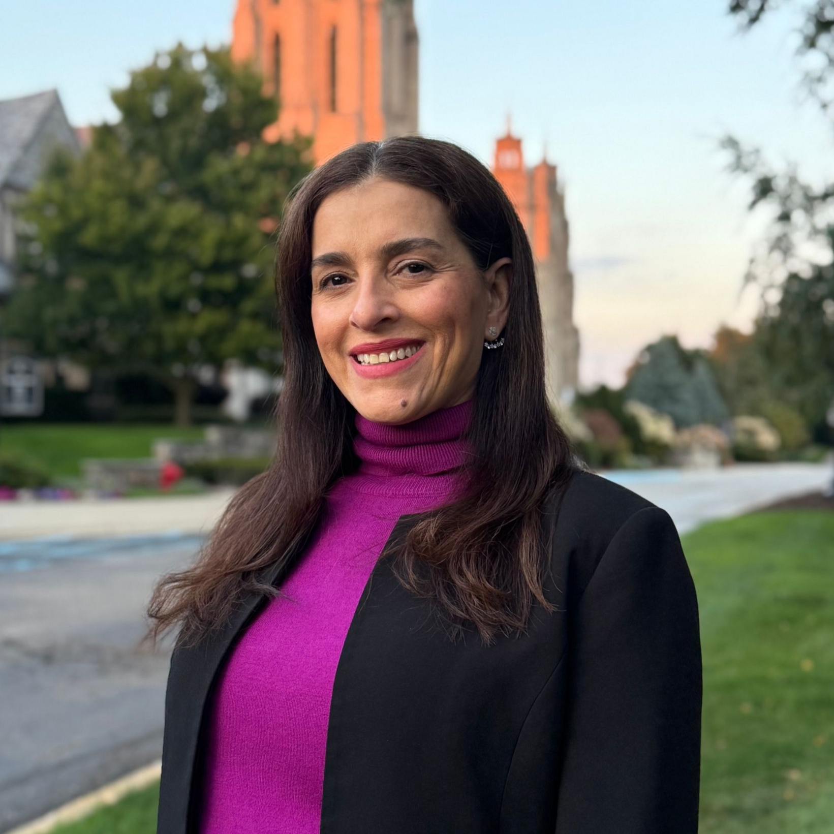 Woman with dark hair in magenta turtleneck and black jacket smiles outdoors, with a building in the background.