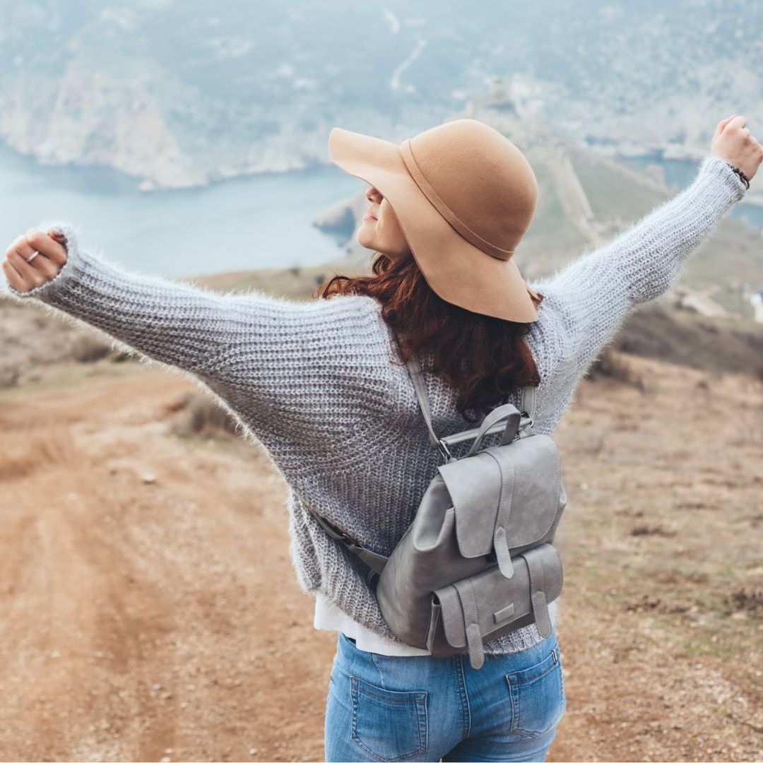 Hope Against Trafficking / A woman wearing a hat and a backpack is standing on top of a hill with her arms outstretched.