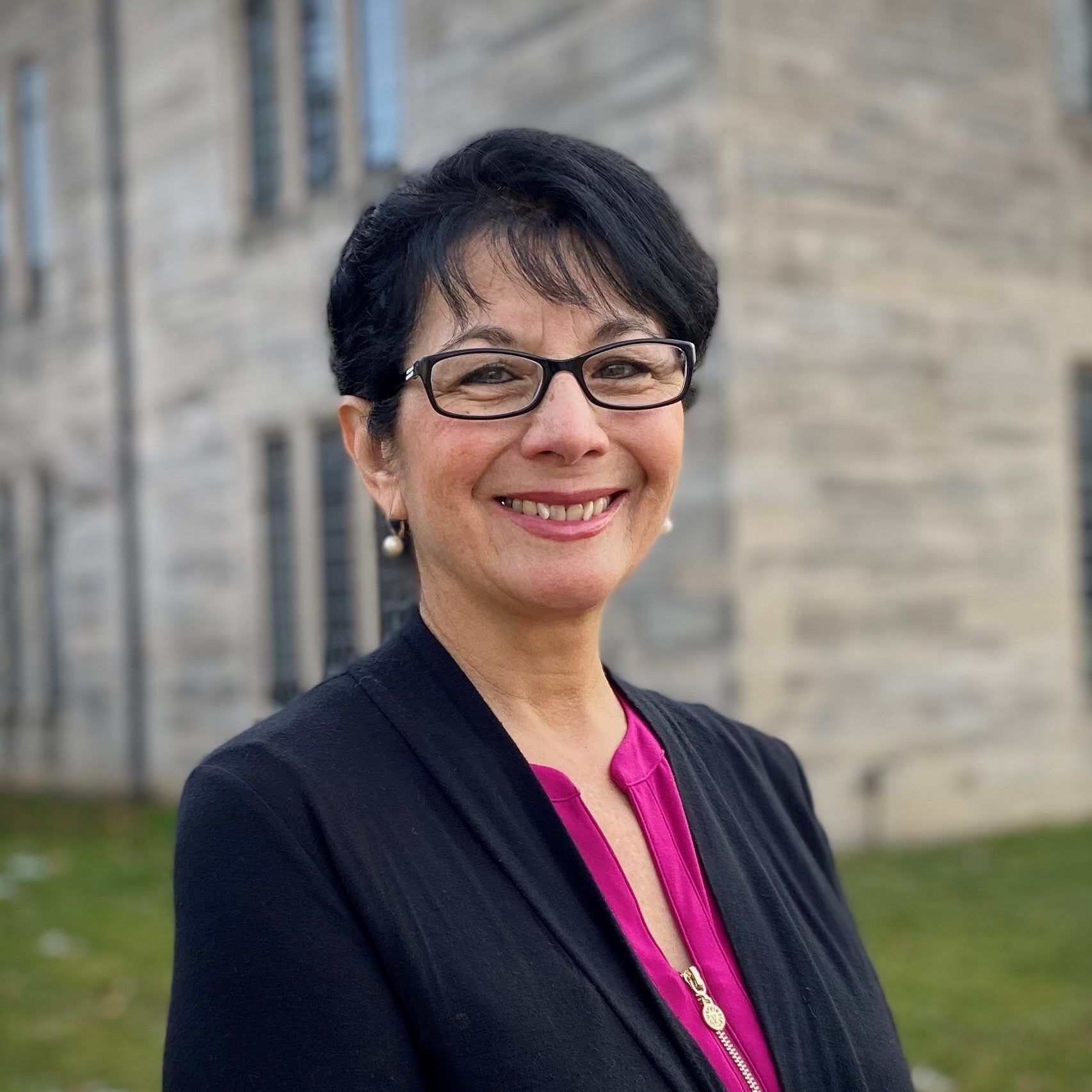 A woman wearing glasses and a black jacket is smiling in front of a building.