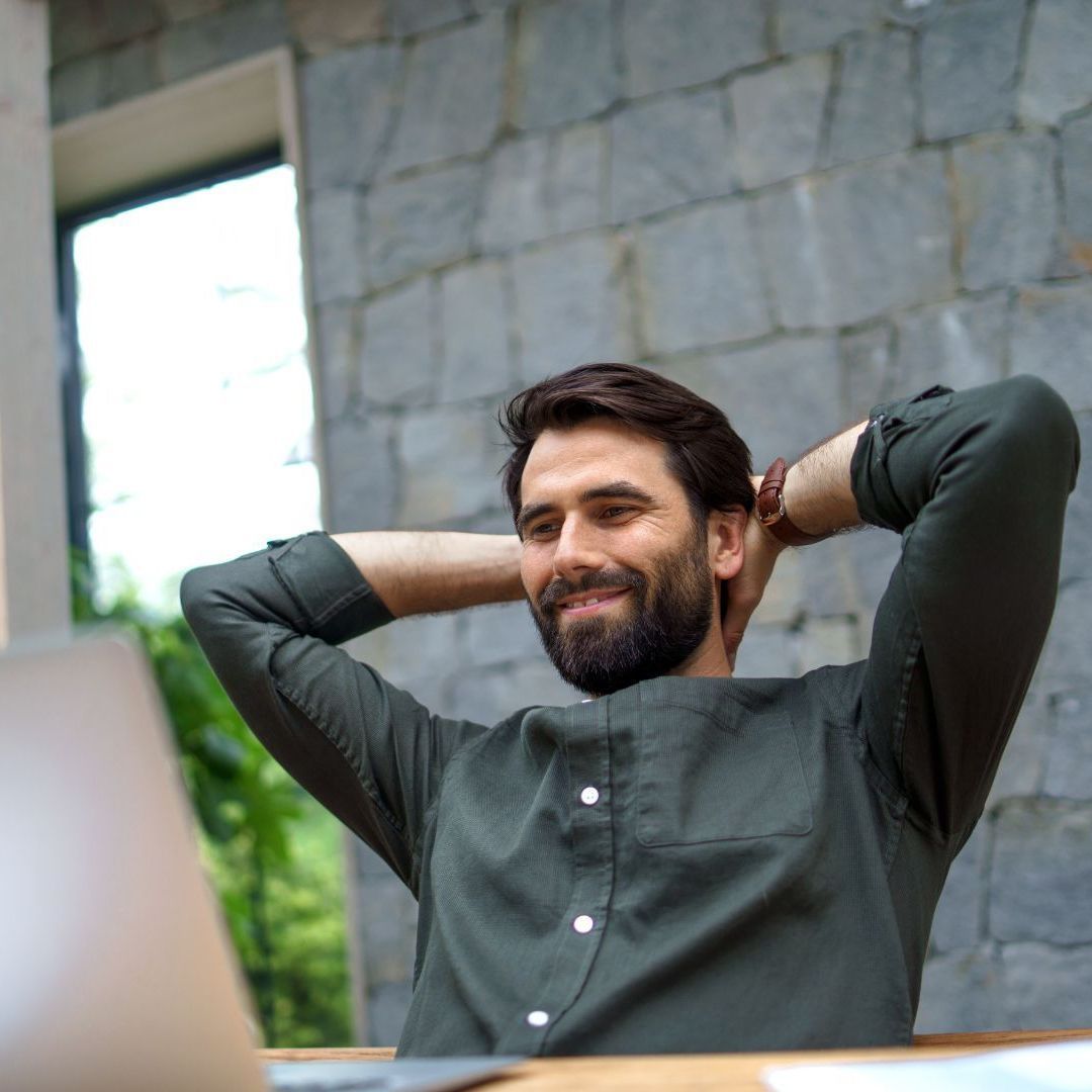 A man with his hands behind his head is sitting in front of a laptop computer.