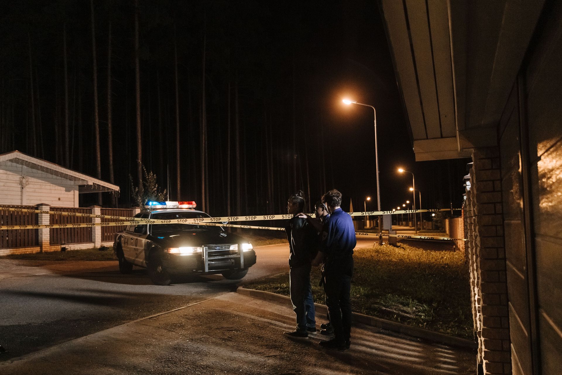 Two police officers arrest a criminal beside a car at night near a building, with streetlights and taped-off area.