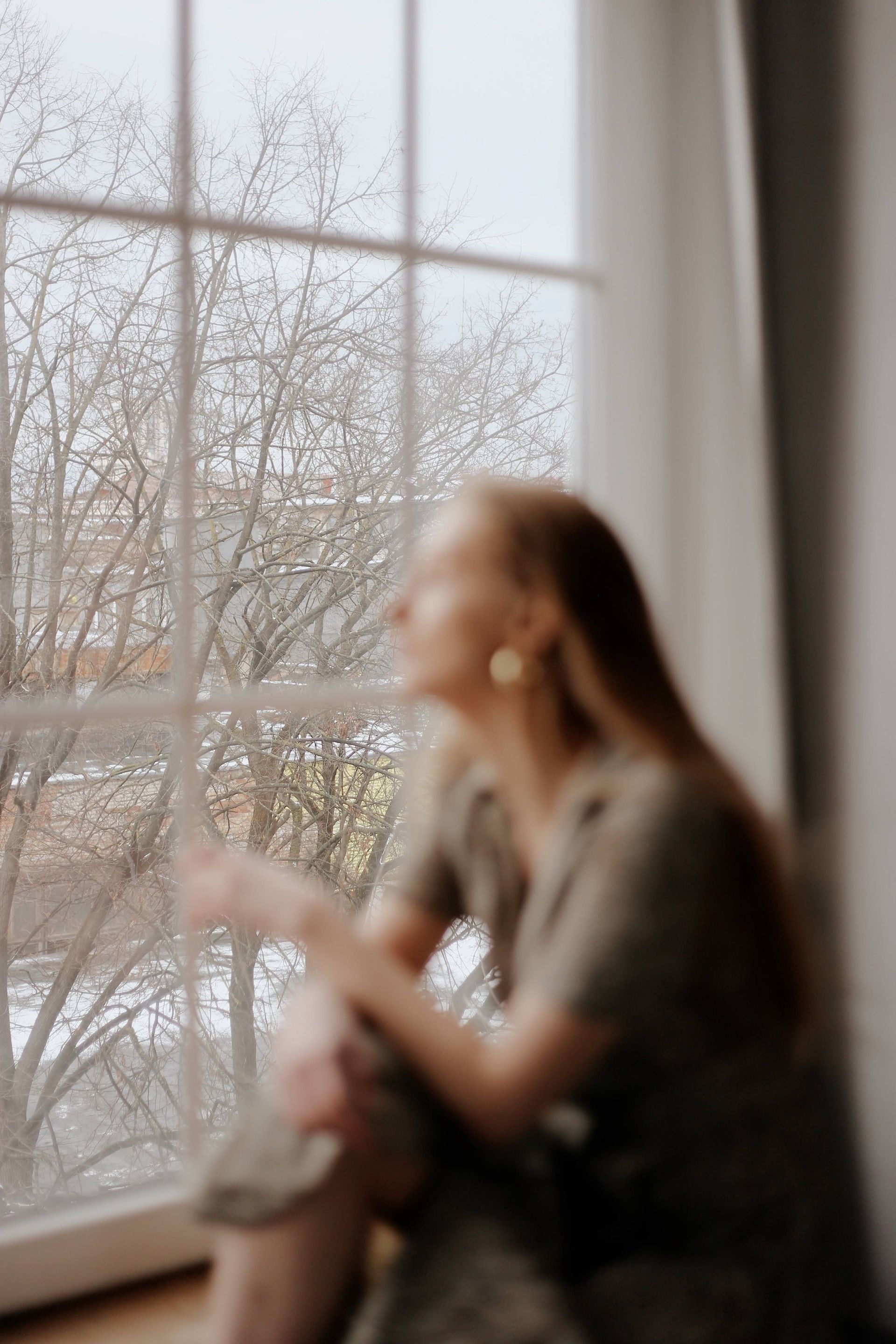 Blurred person sitting by a window, looking outside at bare trees on a gray day