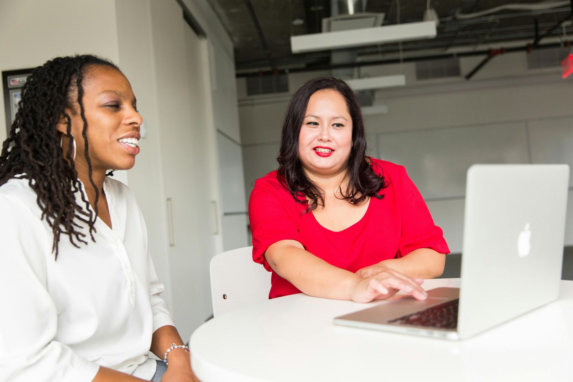 Two women look at laptop at round table; one in white shirt, the other in red, smiling.