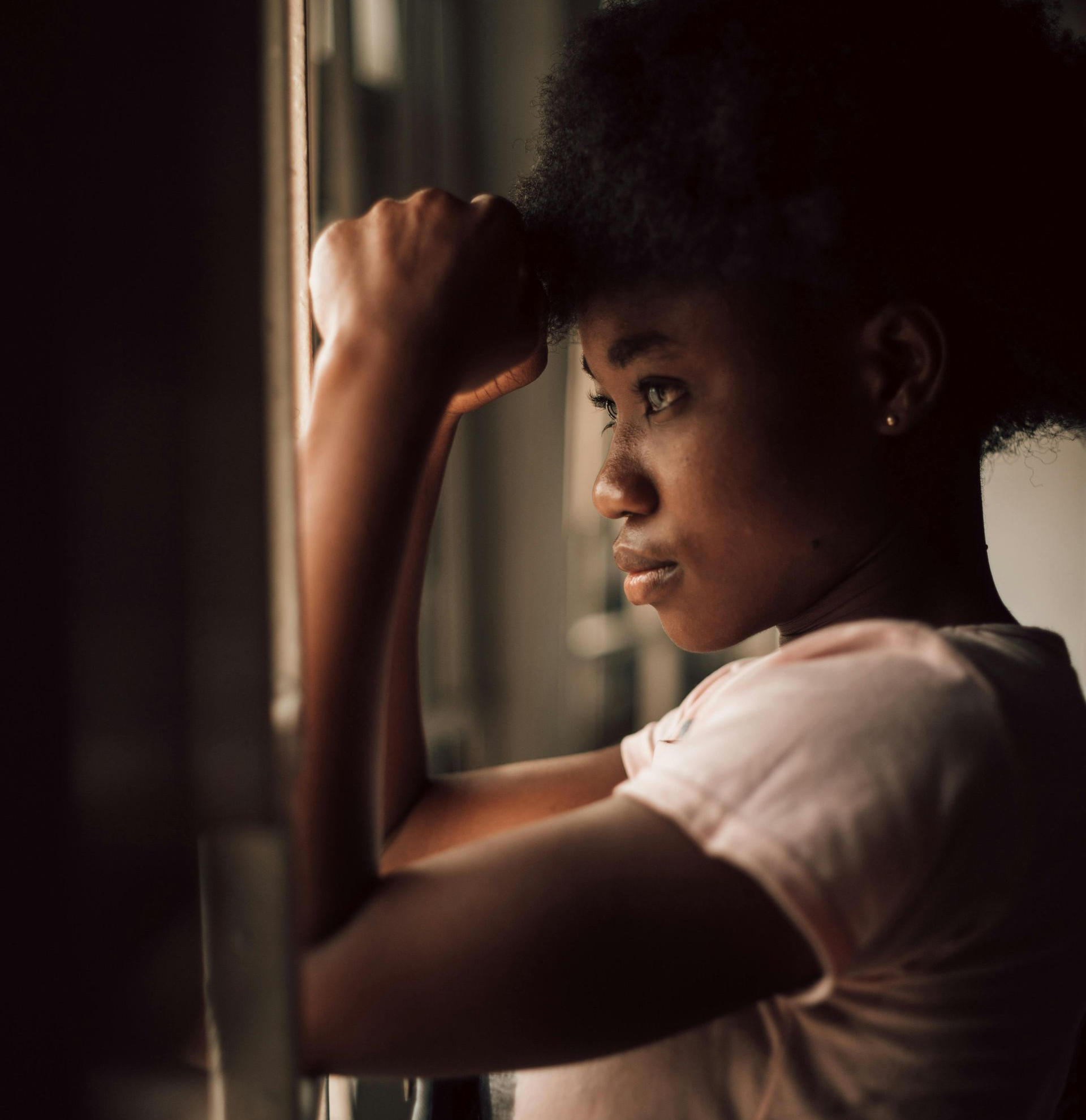Person in a white shirt gazes out a window, resting their arm on the frame in soft light