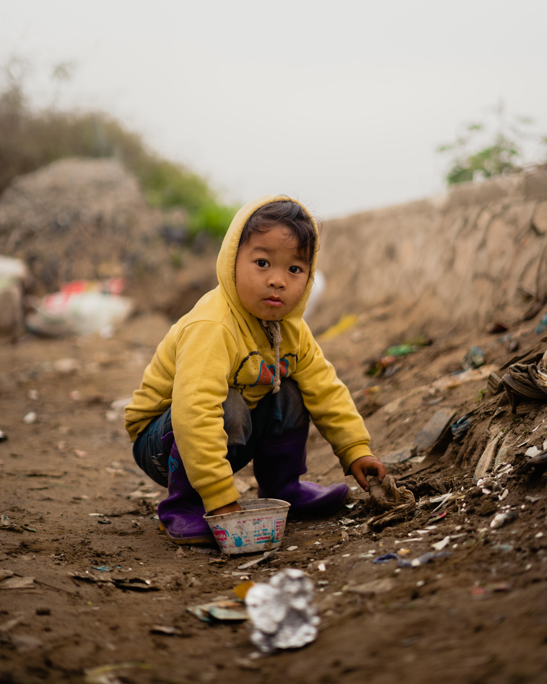 Child in a yellow hoodie crouches on a dirt path, reaching toward litter near a rocky hillside.
