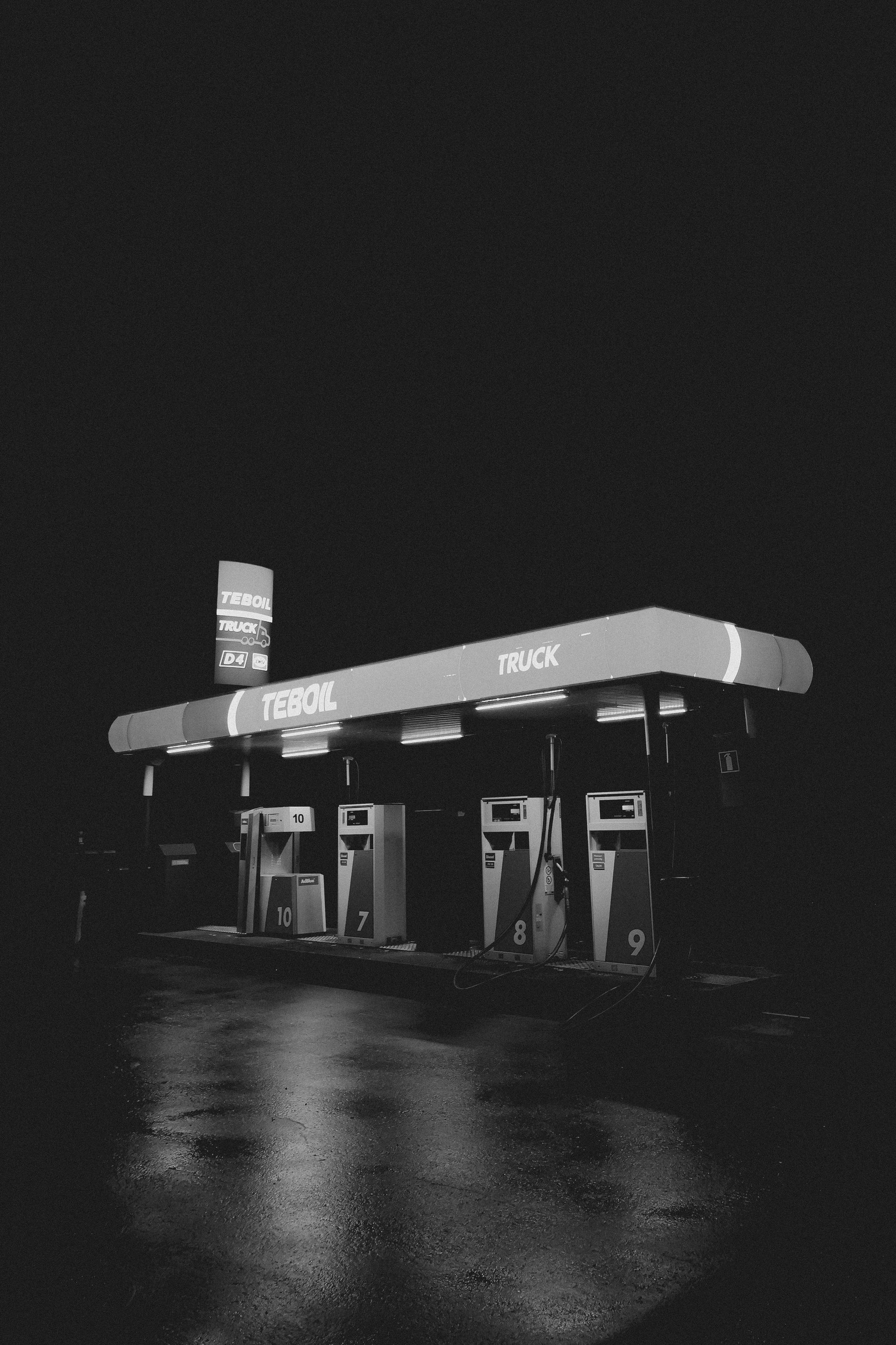 Black and white photo of a gas station at night. Reflective wet ground. Lit signage and pumps.