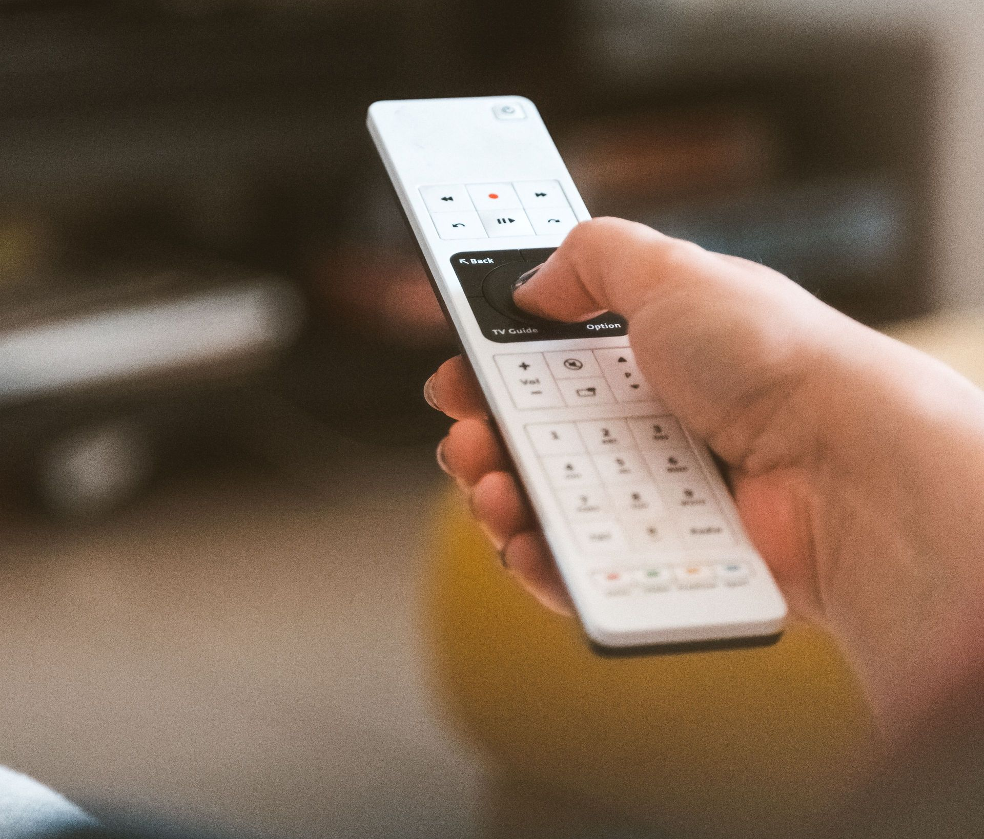 Hand holding a white remote control pointed at a TV in a blurred living room