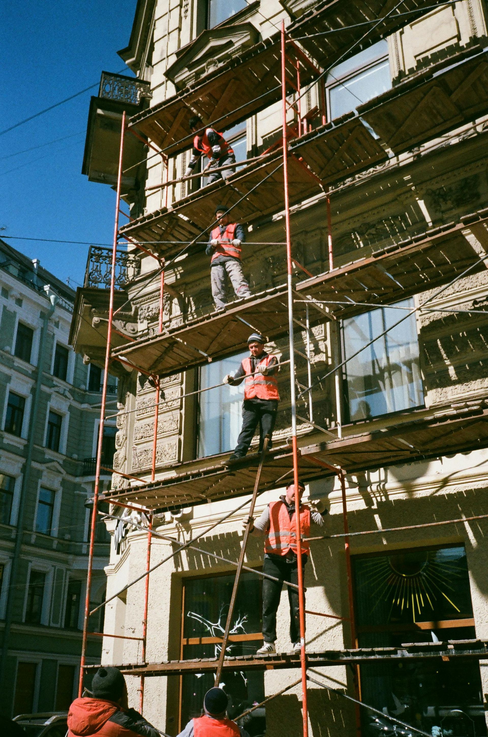 Workers on scaffolding, repairing a building facade. Sunny day. Red vests, safety helmets.