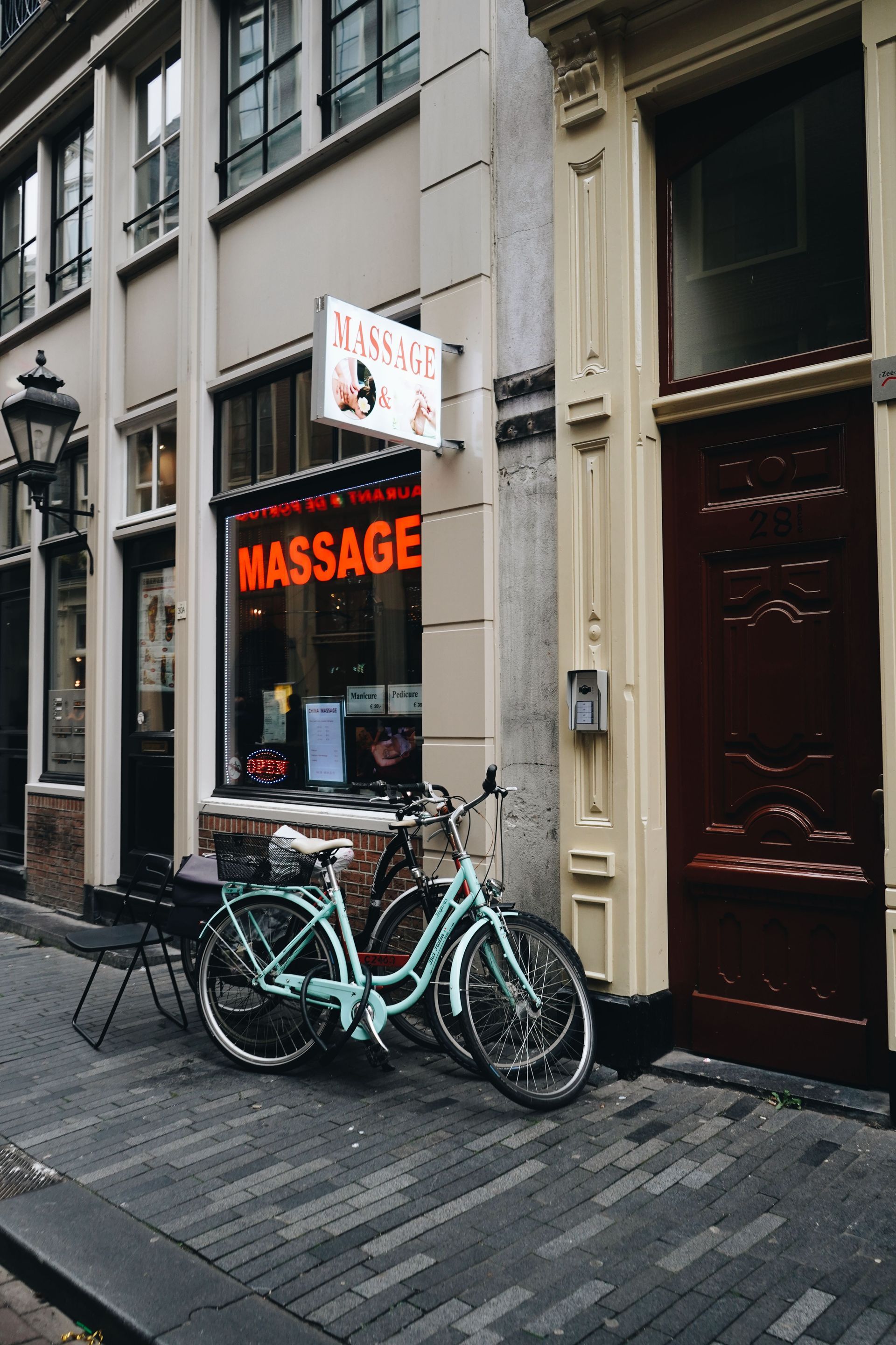 Bicycles parked outside a narrow storefront with a red neon “MASSAGE” sign on a cobblestone street