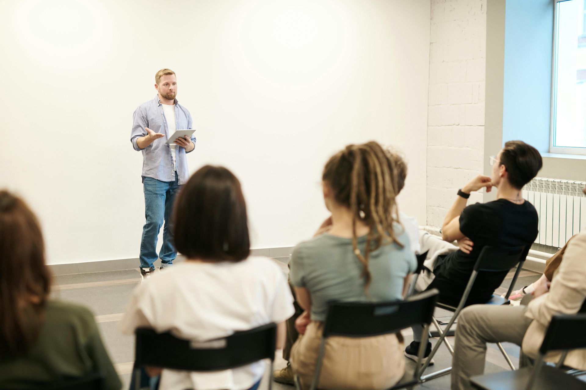 Man presenting to a seated audience in a white-walled room; man holding a tablet.