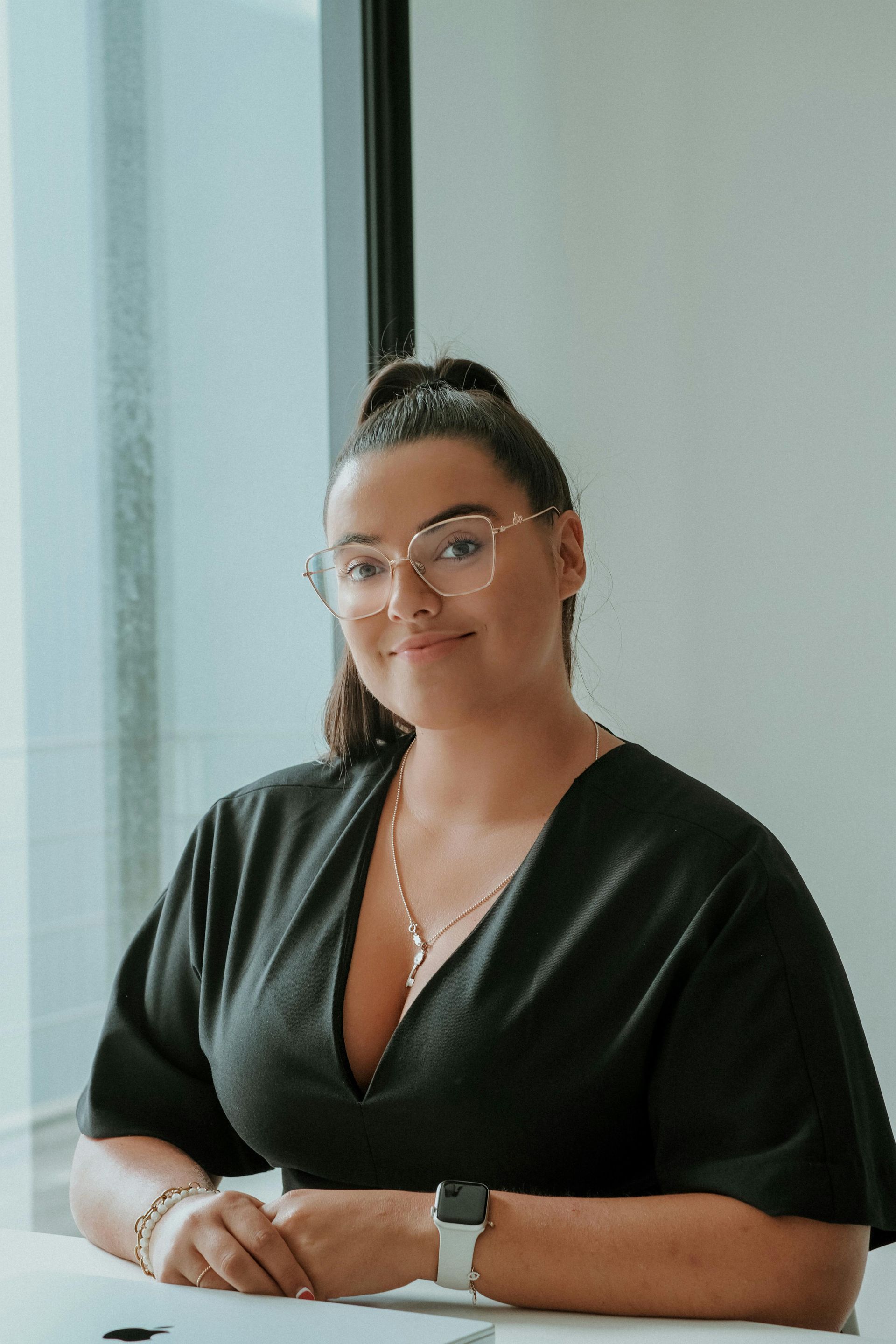 Woman with glasses at a desk, wearing a black shirt, smiling.